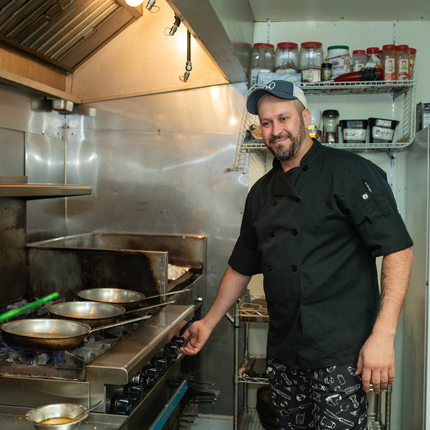 Un hombre de pie frente a una cocina con tres sartenes sobre tres hornillas. El hombre lleva una chaqueta de chef negra y una gorra de béisbol. Hay especias detrás de él en una rejilla y un refrigerador blanco.