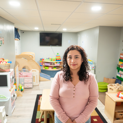 Una mujer con cabello castaño rizado que le llega justo por debajo de los hombros, vestida con una camisa rosa de manga larga, sonríe a la cámara. Está de pie en medio de una sala de juegos infantil con juguetes coloridos que cubren las paredes y una mesa de madera del tamaño de un niño en el centro.