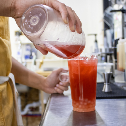 Una taza transparente con un líquido rojizo y perlas de boba de color más claro en el fondo del vaso. El líquido se vierte en el vaso desde otro vaso transparente.  