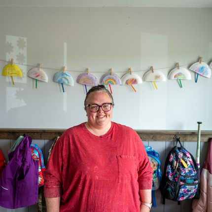 Woman with a 3/4 sleeve dark pink shirt with a pocket, looks at the camera. Her hair is pulled back and she's wearing glasses. She stands in front of a wall of hooks for little one's coats and backpacks. Above the hooks is a string of half paper plate bright constructions.