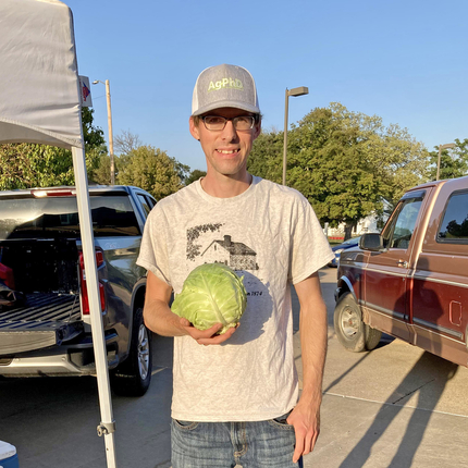 Un hombre con gorra de béisbol, camiseta y vaqueros sostiene una lechuga y mira a la cámara. Al fondo se ven las partes traseras de dos camionetas y la pata de una tienda de campaña.
