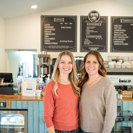 Dos mujeres frente a un mostrador donde pides café, con un tablero de menú detrás de ellas