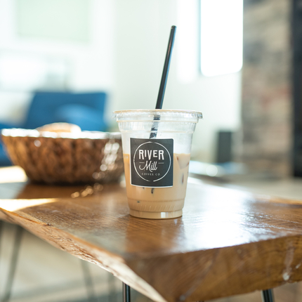 Plastic cup fill with beverage with River Mill logo sticker and straw in black on a wooden table