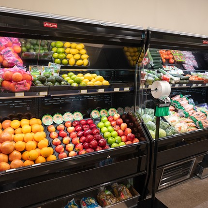 Photo of a fruit and vegetable open cooler at a rural grocery store with oranges, red and green apples, lemons, bags of carrots, heads of lettuce.
