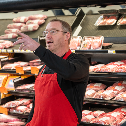 A man stands in front of a grocery store meat cooler with his left hand raised, appearing to be speaking. He wears glasses, a black long sleeve shirt, and a red apron. 