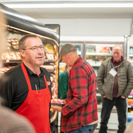 Man with short, brown hair and glasses stands in front of a grocery cooler wearing a black polo shirt and a red apron over it.