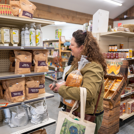 Woman with curly hair, wearing a coat, holds a loaf of bread and a tote bag. She's in a grocery store looking at a shelf of flour and oils.