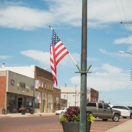An American flag waves, suspended from a weathered green post on a rural main street. In the background, we see vehicles parked on a brick road and historic brick-front buildings. There is a potted plant below the flag and the sky is bright blue with wispy clouds.