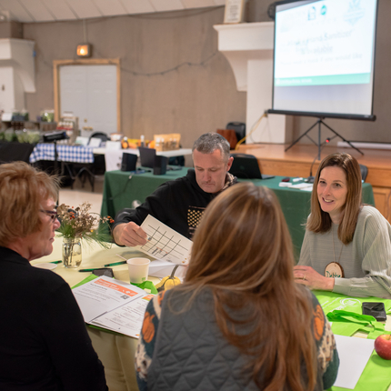 Three white women and a white male sit at a long table with papers, fruit, and other items on the table. A large white projection screens is in the background near a stage and an exit door.