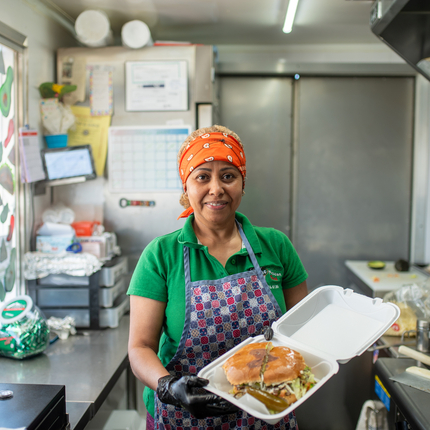 Latina woman wearing a red bandana, green shirt, and purple and pink checkered apron holds an open styrofoam container with a large sandwich inside. She's standing inside a food truck with the window to the left of the photo.