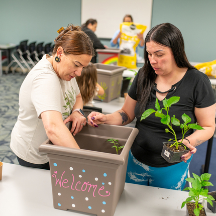 Dos mujeres nativas americanas colocan plantas en una maceta cuadrada con tierra que dice "bienvenida"; la maceta está sobre una mesa en un aula alfombrada