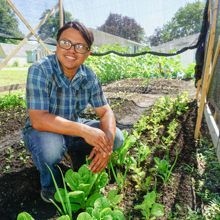 Hombre latino con gafas, camisa a cuadros azul de manga corta y jeans, arrodillado entre hileras de verduras