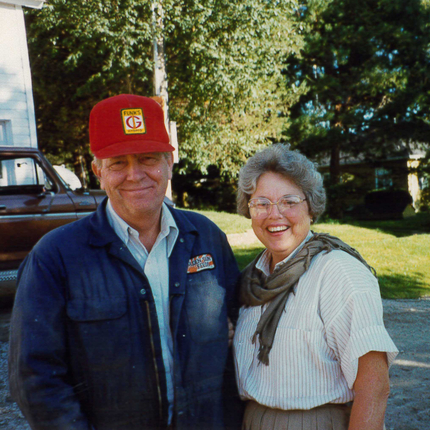 La foto es de los años 1980. Un hombre y una mujer, de unos 60 años, sonríen a la cámara. El hombre lleva una gorra roja y una chaqueta azul. La mujer tiene el pelo corto y canoso, una camisa blanca y una bufanda verde. Detrás de ellos se ve una calle rural, un patio con pinos y un gato cruzando la calle.
