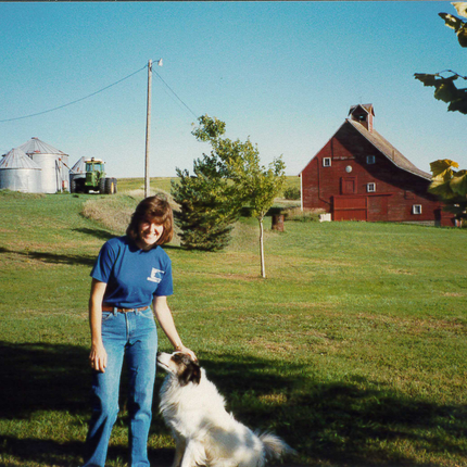 Una mujer con cabello castaño hasta los hombros, con camiseta azul y vaqueros, sostiene en una mano a un perro blanco y peludo con la cara oscura. Está de pie en un patio con un granero y silos al fondo. La foto es de la década de 1980.