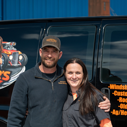 A woman and a man stand in front of a black van. On the van is an image that says Schroyer's Mobile Flat Glass, windshield replacement, custom cut flat glass, rock chip repair, oldies/hot rods, ag/heavy eq. The man is wearing a dark colored fleece with a collar and a ball cap. The woman has long straight brown hair and is wearing a button up short sleeve shirt.