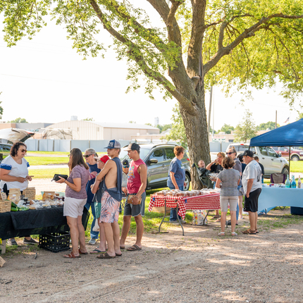A row of farmers market vendors along a road. Vendors have tables with bright colored clothes and products on top. Groups of customers are in front of each table.