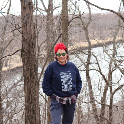 A woman with short, bright red and black hair stands in a leafless patch of trees near a river. They are wearing reflective sunglasses, a navy blue sweatshirt with a diagram labeled "Power Stroke," a plaid shirt underneath, and gray cargo pants. Their hands are in their pockets.