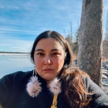 Portrait from the shoulders up of a woman with long dark hair wearing a black coat and fluffy pom‑pom earrings, standing beside a tree on a calm lakeshore under a clear blue sky.