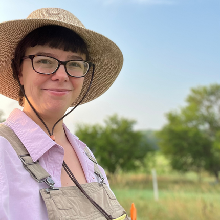 Woman with short dark hair, a sunhat, and dark glasses, looking at the camera. She is also wearing a collared pink shirt and beige overalls. In the background is a blue sky and green trees and green grass.