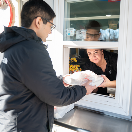 Woman in a black polo smiles as she hands a meal through a white food truck window to a man in a black jacket.