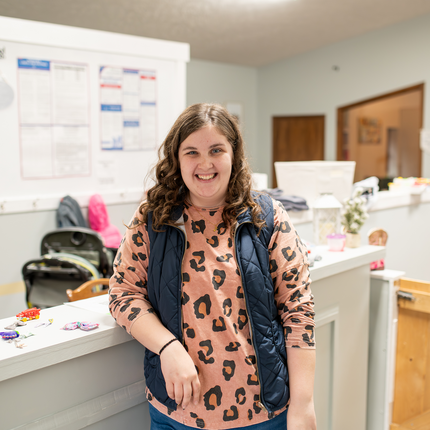 A woman with shoulder-length curly brown hair leans against a white countertop. She wears an orange leopard-print shirt with a black vest.
