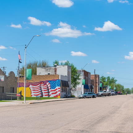 Rural Main Street, with a big flag mural on the side of a building on the left side of the street. About two blocks of downtown buildings with cars parked on both sides