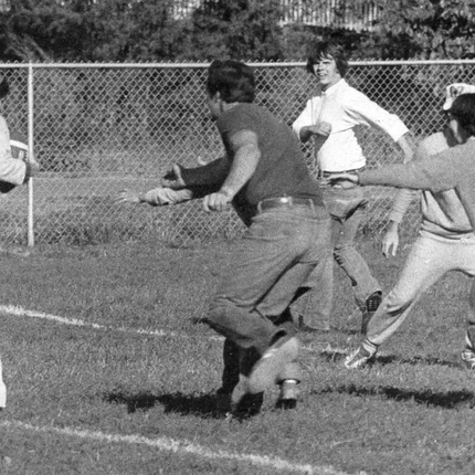 Una fotografía en blanco y negro de los años 70 de adultos jugando al fútbol de toque. Hay 2 mujeres con camisas blancas y un hombre y una mujer vestidos de gris arremetiendo hacia la dama de blanco con la pelota.