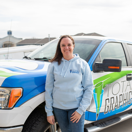 Una mujer blanca con cabello castaño hasta los hombros y vestida con una sudadera con capucha azul claro se para frente a un camión con diseños azules, blancos y verdes y las palabras "Total Graphics" en el lado del conductor.