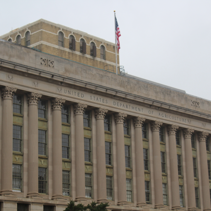 Edificio del USDA en Washington, D.C. Es un edificio grande de varias plantas con intrincadas columnas y ventanas en la fachada. En la parte superior, se puede leer el lema "UNITED STATES DEPARTMENT OF AGRICULTURE" (Departamento de Agricultura de los Estados Unidos). La bandera estadounidense ondea en un asta en el tejado.