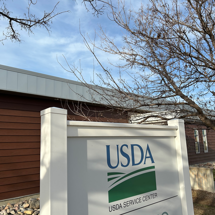 Un letrero blanco con las palabras "USDA SERVICE CENTER" y el logotipo verde de las colinas onduladas. Al fondo, un edificio de una sola planta con revestimiento marrón rojizo.