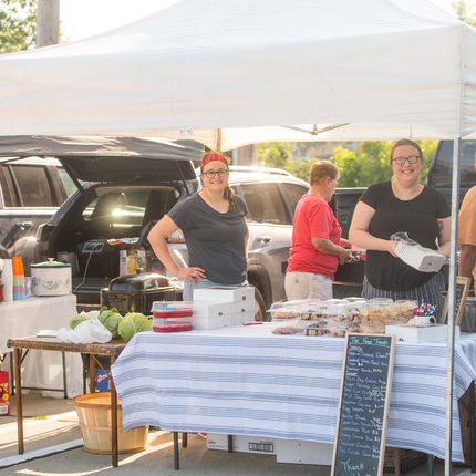 Two women standing at a table with baked goods with a tent over them at a farmers market