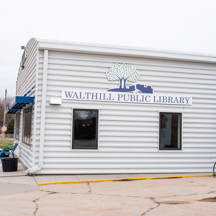 A large building with horizontal white metal siding. There are two windows with a blue sign above that says "WALTHILL PUBLIC LIBRARY". The sign also includes a silhouette of trees and a cityscape. Around the corner of the building, we see two blue awnings overhanging two sets of windows and a blue metal bench with black planters on both sides of it.