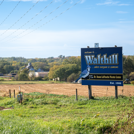 A roadside welcome sign for Walthill, Nebraska, stands beside a grassy field under a clear blue sky. The sign reads “Welcome to Walthill… where everyone is someone,” and notes it is the home of the Dr. Susan LaFlesche Picotte Center and established in 1906. Behind the sign are harvested fields, scattered trees, and a few buildings in the distance.