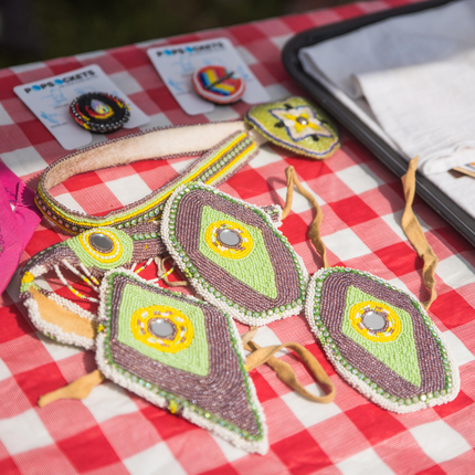 A close-up photo of a red and white checkered table covered with handmade beaded jewelry and accessories, including geometric-patterned pieces with green, yellow, and brown beads, circular pins on display cards, and colorful embroidered patches. The items are arranged on the table in bright sunlight.