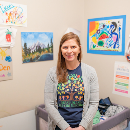 A woman with light brown hair stands in her day care looking directly at the camera smiling. The walls are decorated with bright colored pictures drawn by children.