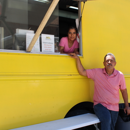 A man stands in front of a bright yellow food truck and a woman looks out from the food truck window