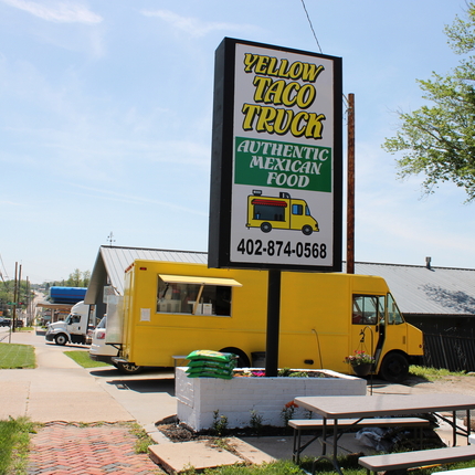 A view from down the sidewalk toward a bright yellow food truck and a permanent sign that says "Yellow Taco Truck"