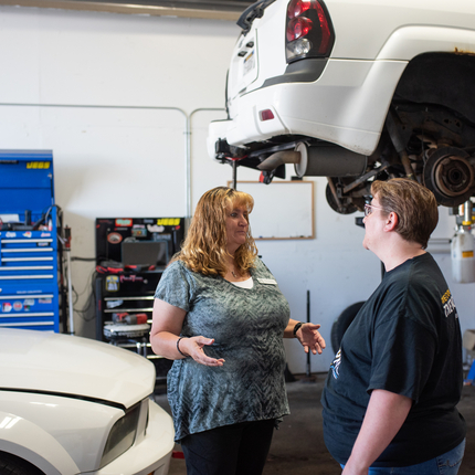 Dos mujeres de pie en un taller de reparación de automóviles, con un automóvil levantado sin las ruedas