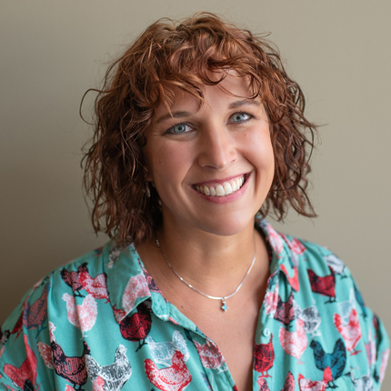 White woman with short red curly hair and blue eyes, wearing a teal shirt with chicken designs in read, pink and grey colors smiles at the camera