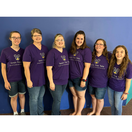 Six women wearing purple shirts and blue jeans and shorts stand in front of a lavender wall posing for a picture