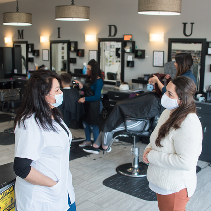 Two women with masks face to face at a beauty salon, with two women doing people's hair behind them