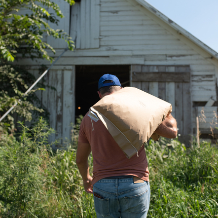 Man carrying a sack of feed with grass in the foreground and barn in the distance.