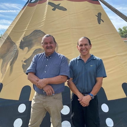 two Native American men wearing blue polos standing in front of a beige teepee with buffalo and birds on it