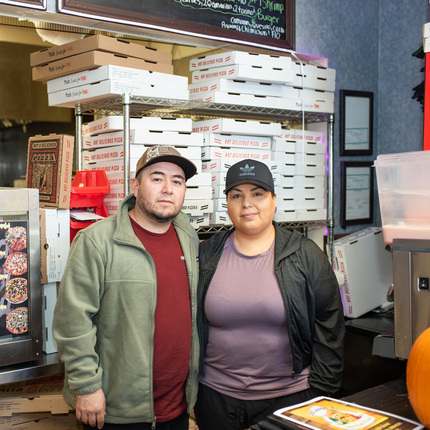 A man and a woman stand in front of a pile of pizza boxes behind them in a pizza shop