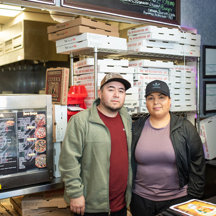 Un hombre y una mujer posan para una foto en frente de una repisa con cajas de pizza