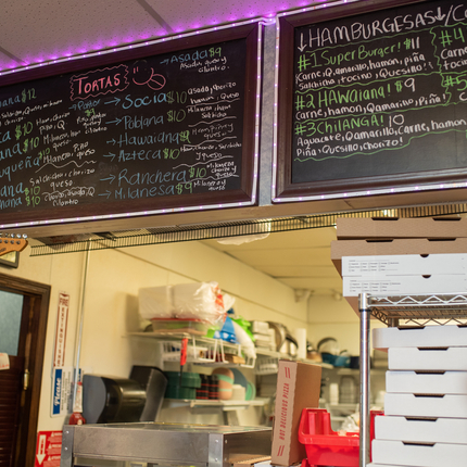 Black menu with food options listed in colorful writing above a shelf with pizza boxes