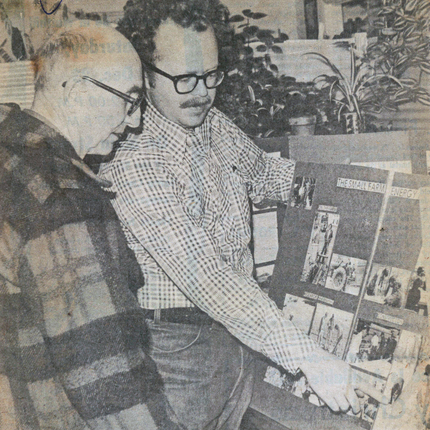Foto de periódico antiguo (de los años 1970) de dos hombres. Un hombre con un abrigo a cuadros mira a otro hombre con una camisa de manga larga con botones a cuadros y señala una cartulina con fotos.