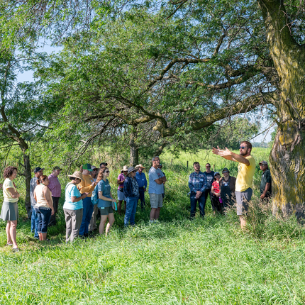 A large group of people standing in a field, under a tree. A man in a yellow t-shirt and sunglasses appears to be addressing the crowd with his arms stretched out in front of him.
