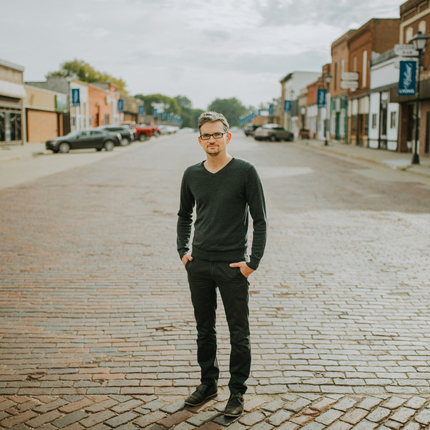 Man in dark gray shirt and black pants standing in the middle of a brick street in a small town, with 2-story buildings on either side of the street
