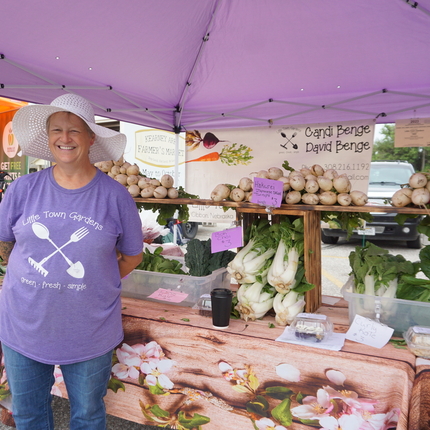 Woman in t-shirt that says "Little Town Gardens" in front of a vegetable stand  with bok choy, onions, and more